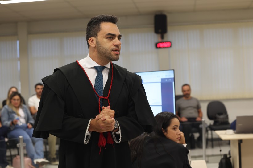 Homem de terno e capa, falando em uma sala de aula, com pessoas assistindo ao fundo.