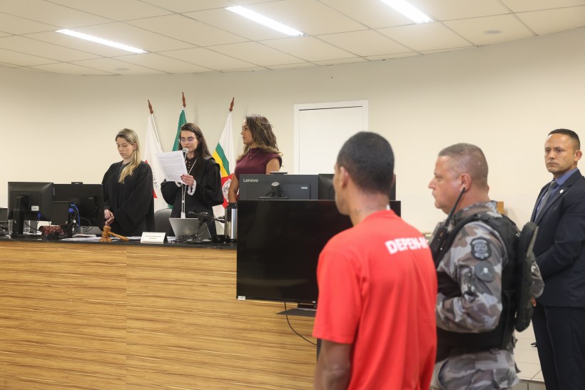 Audiência judicial com juízas, advogados e réu em uniforme vermelho, em um tribunal.