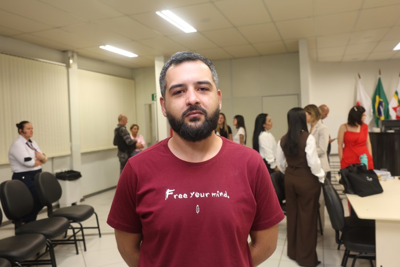 Homem com barba e cabelo curto, vestindo uma camiseta vermelha com a frase 'Free your mind', em ambiente com outras pessoas ao fundo.