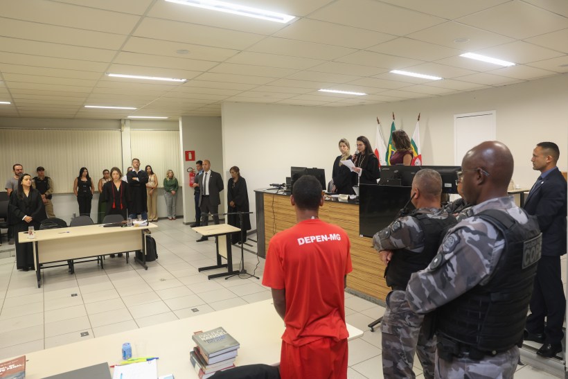 Audiência judicial com réu em uniforme vermelho, juízes e advogados em pé na sala de tribunal.