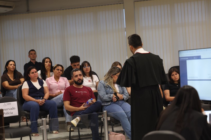 Um grupo de pessoas assistindo a uma apresentação em um ambiente formal, com um apresentador vestido com uma túnica na frente, enquanto um monitor exibe informações.