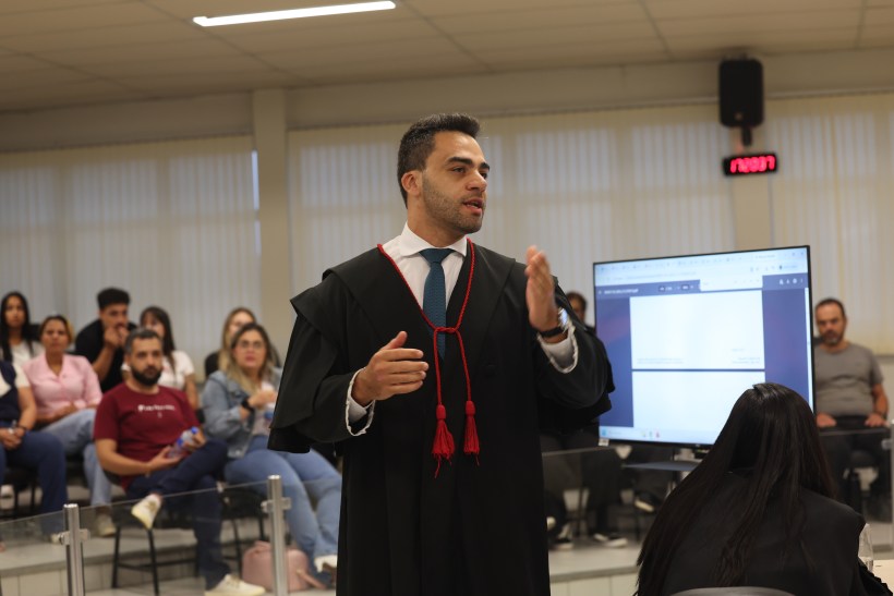Homem em traje acadêmico falando em uma sala de aula cheia de alunos, com um projetor ao fundo.
