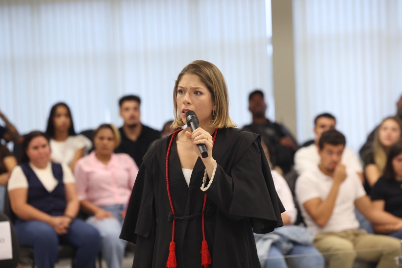 Mulher em traje jurídico falando ao microfone diante de uma audiência, com público ao fundo.