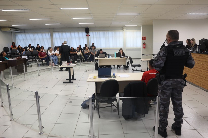 Sala de audiência com várias pessoas sentadas, incluindo um juiz em pé, um policial com uniforme tático e pessoas na bancada do tribunal. Há mesas com equipamentos e documentos.