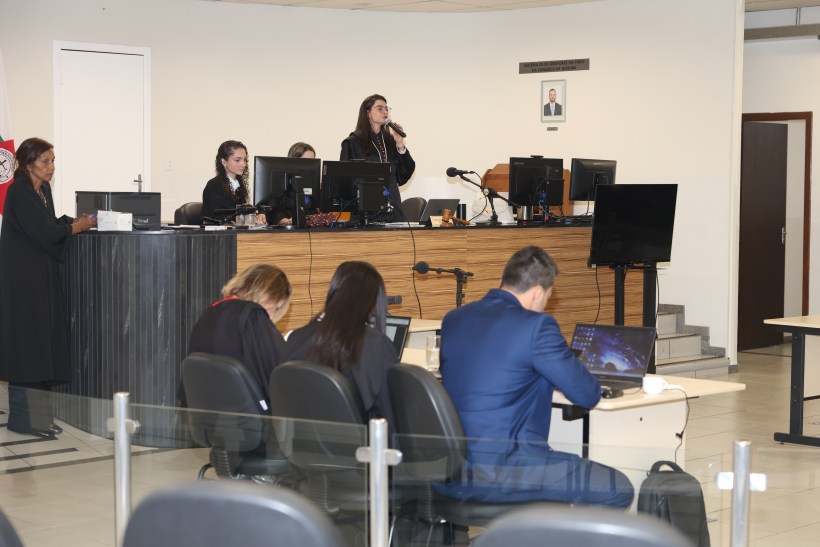 Ambiente de tribunal com pessoas em atividade. Uma mulher fala ao microfone no púlpito, enquanto outras pessoas trabalham em computadores e um advogado parece escutar atentamente.