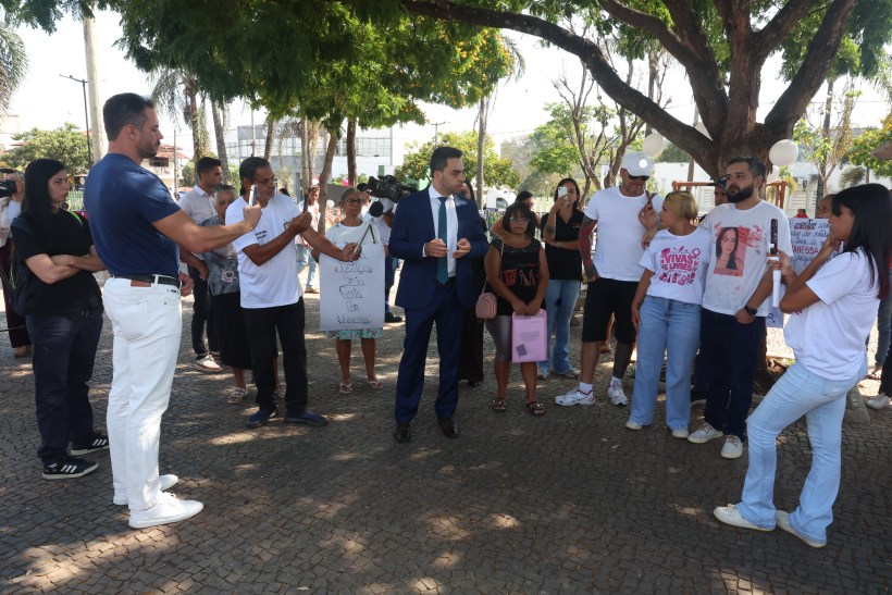 Grupo de pessoas em um protesto ao ar livre, com cartazes na mão e interagindo com um homem em traje formal.