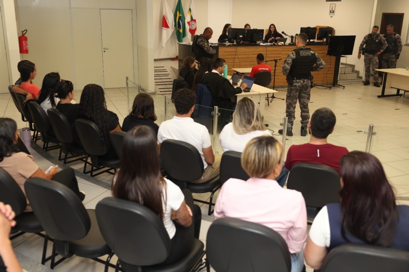 Audiência judicial com juízes, advogados e policiais em tribunal. Vários espectadores assistindo.