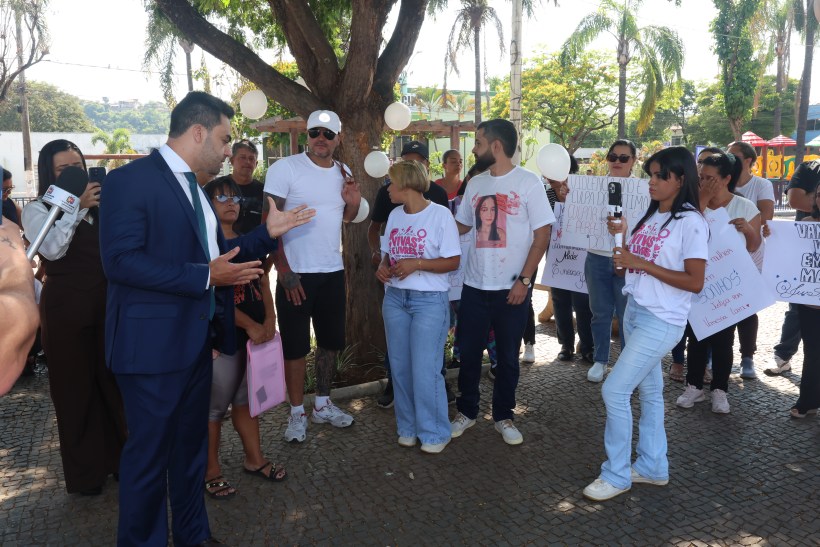 Grupo de pessoas em protesto; alguns seguram cartazes e balões enquanto um homem de terno e gravata fala com elas sob uma árvore.