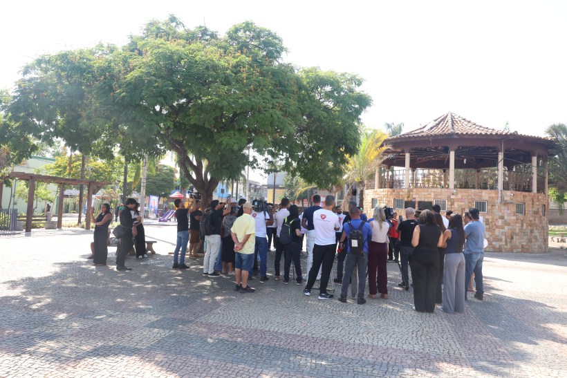 Grupo de pessoas se reunindo em uma praça, com um grande árvore ao fundo e um quiosque visível.