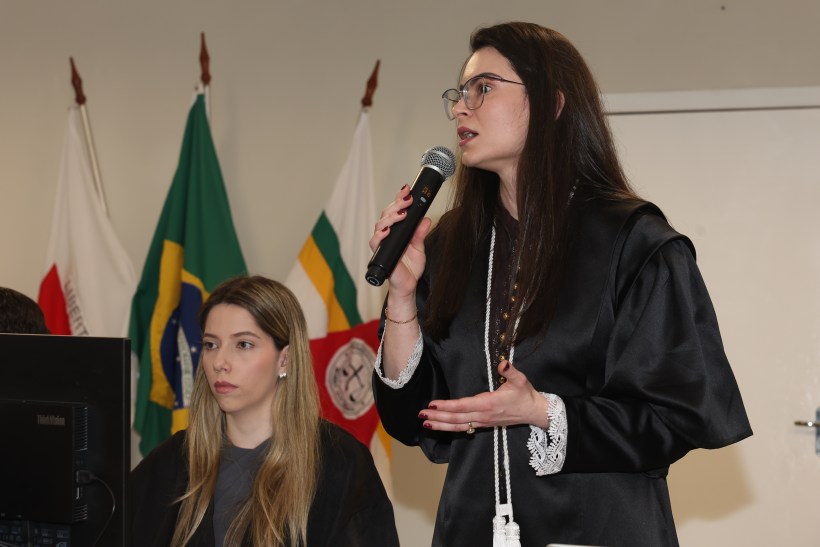Uma mulher em trajes formais segurando um microfone enquanto fala, com outra mulher sentada ao lado e bandeiras ao fundo.
