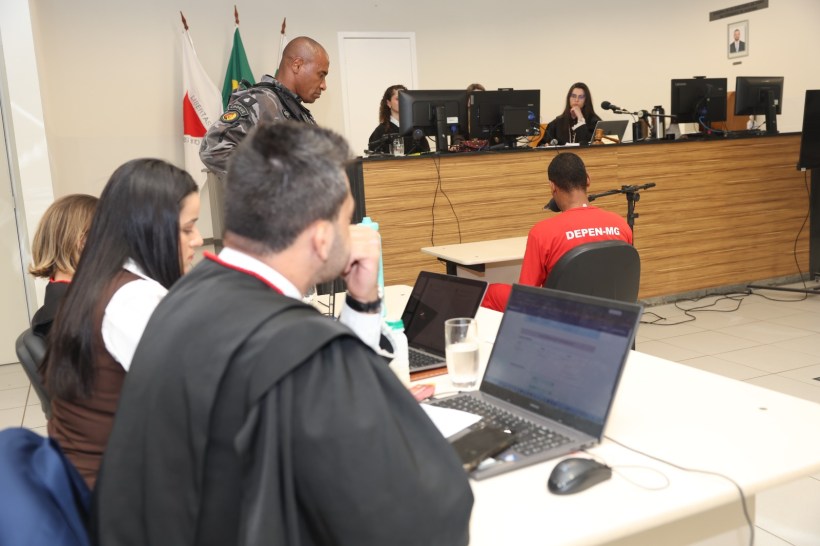 Um tribunal com várias pessoas, incluindo um policial e um réu vestido de vermelho, na frente de computadores e mesas.