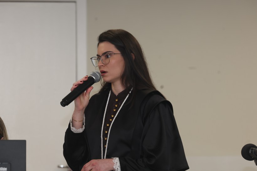 Mulher com óculos de grau, vestindo um traje formal preto, segurando um microfone enquanto fala em um evento.