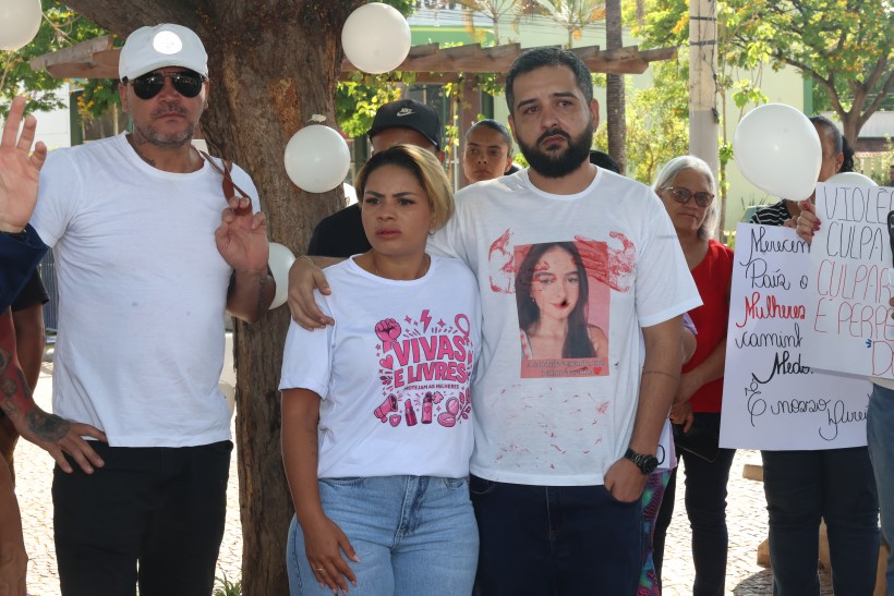 Grupo de pessoas em protesto, com foco em mulheres e direitos humanos, algumas vestindo camisetas com mensagens de apoio. Ao fundo, cartazes e balões brancos.