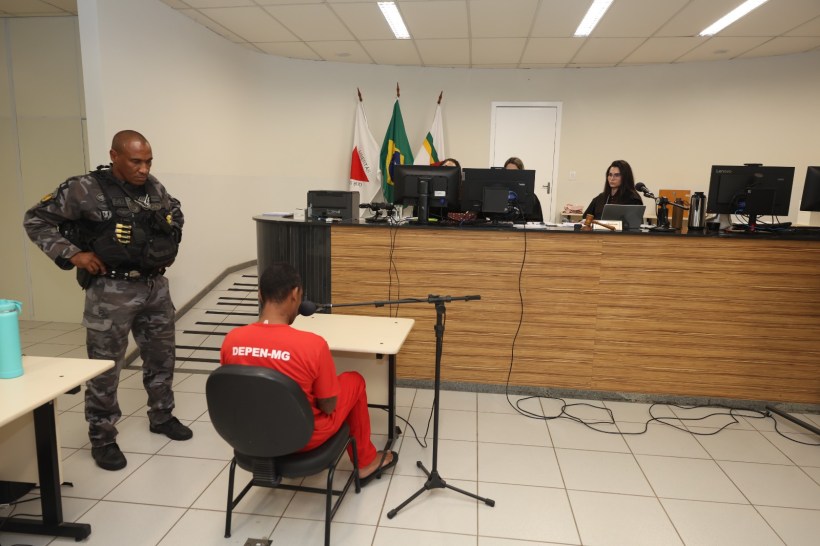 Homem sentado em uma cadeira, vestido com um uniforme vermelho, diante de um tribunal com computadoras e três pessoas observando. Um policial militar em pé ao lado.