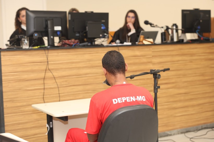 Homem sentado em uma cadeira, vestindo uniforme vermelho com a identificação 'DEPEN-MG', diante de uma bancada com profissionais da justiça em um tribunal.