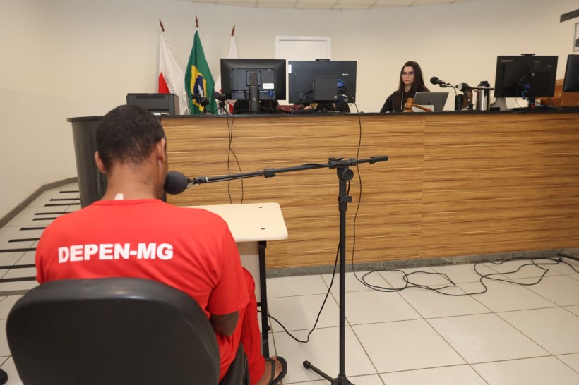 Audiência judicial com um homem usando uniforme vermelho, sentado de costas, e uma profissional ao fundo em frente a computadores.