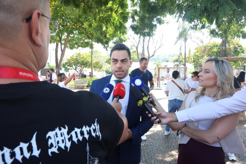 Um homem em terno sendo entrevistado por jornalistas com microfones enquanto outras pessoas estão ao fundo.