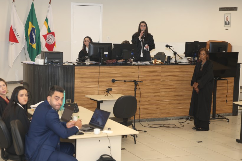 Reunião judicial em tribunal com juízes e advogados. Várias pessoas estão sentadas em mesas, com computadores e documentos, enquanto uma juíza fala ao microfone. Bandeiras do Brasil e de outros estados estão ao fundo.
