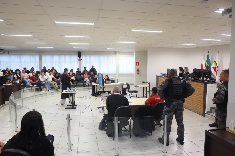 Interior de um tribunal com várias pessoas na audiência. Uma mulher apresenta um caso em pé, enquanto os juízes estão ao fundo. Há segurança armada e um público assistindo.