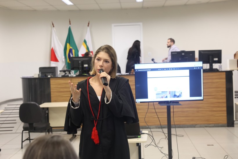 Mulher em toga discursando em um ambiente de sala de aula, com um microfone na mão. Ao fundo, bandeiras do Brasil e de estados, além de computadores em uma mesa.