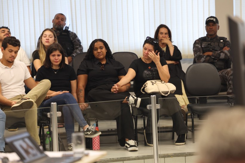 Grupo de pessoas sentadas em cadeira em um ambiente interno, com expressões de tristeza e preocupação. Mulheres vestindo roupas escuras, uma delas enxugando lágrimas. Um policial visível ao fundo.