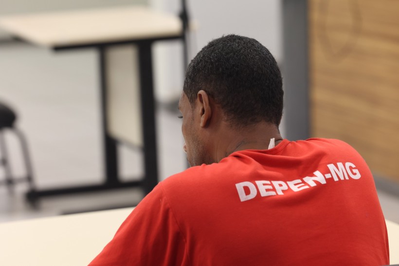 Homem de costas usando camiseta vermelha com a inscrição 'DEPEN-MG', sentado em mesa em ambiente interno.