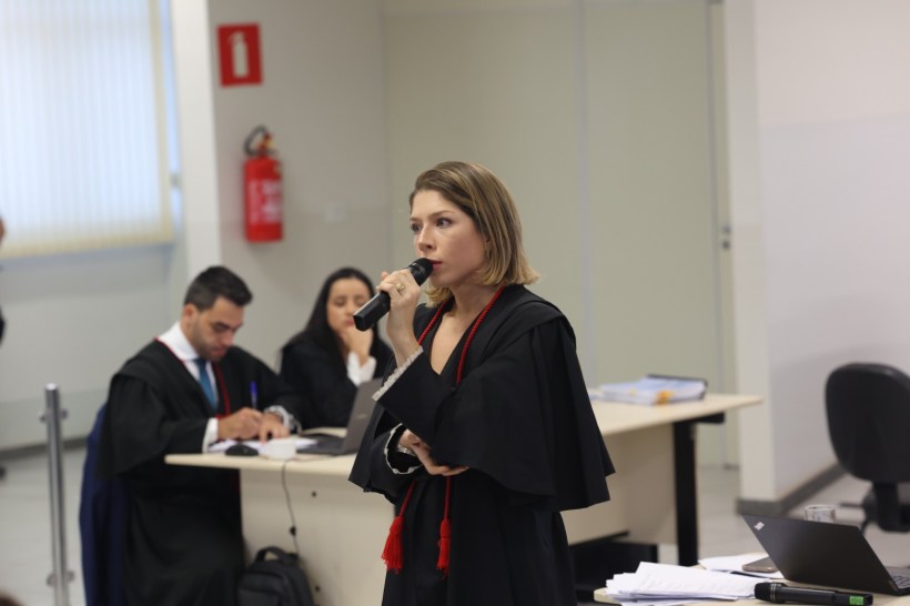 Advogada em pé falando ao microfone, vestida com toga e fita vermelha, durante audiência judicial. No fundo, outras pessoas em posição de discussão e um computador na mesa.