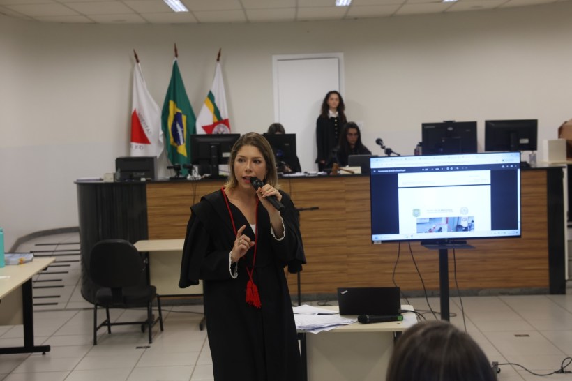 Mulher em toga apresentando em sala de aula com computadores ao fundo e bandeiras penduradas.