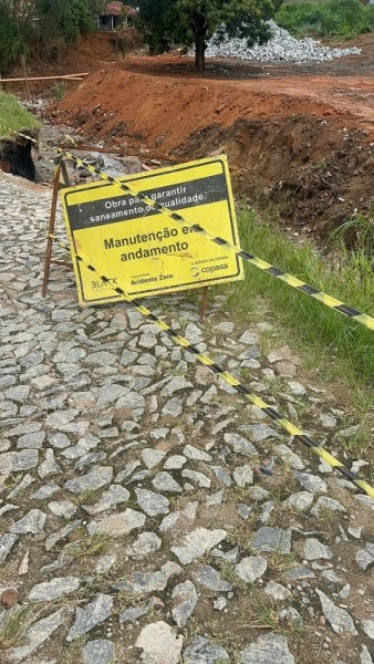 Placa de sinalização em obra de saneamento indicando manutenção em andamento, com fundo amarelo e faixa de segurança preta e amarela.