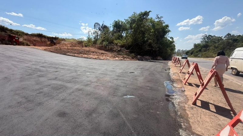 Imagem de uma estrada em construção, com asfalto novo e barricadas de segurança ao lado. Ao fundo, vegetação e um veículo em movimento.