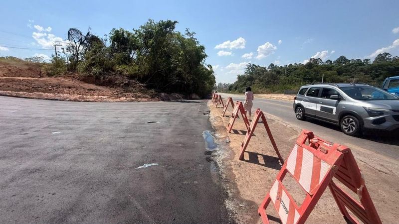 Imagem de uma estrada em obras com barreiras de segurança vermelhas e brancas ao longo do caminho. Ao fundo, veículos passando e árvores ao lado da via.