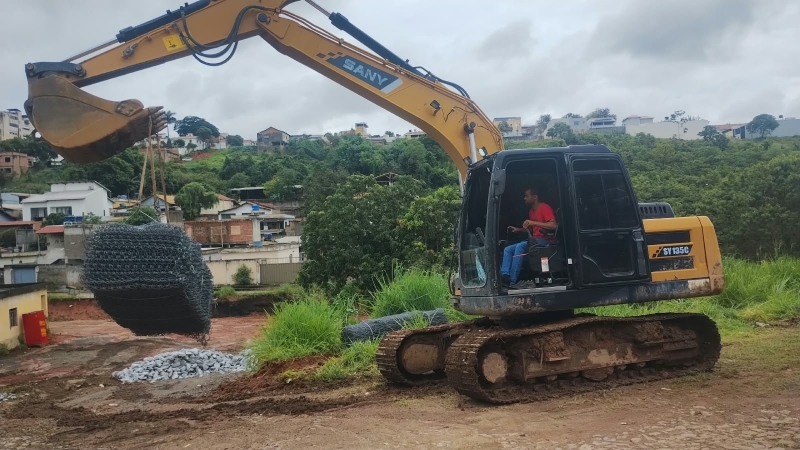 Máquina escavadeira operada por um homem levantando uma grande estrutura em uma área de construção cercada por vegetação e casas ao fundo.