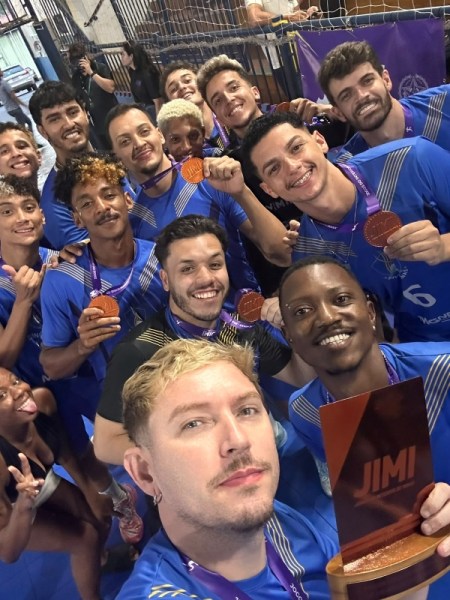 Grupo de jovens atletas sorrindo e comemorando, vestindo uniformes azuis e segurando medalhas de bronze. Um dos membros está tirando um selfie com um troféu na mão.