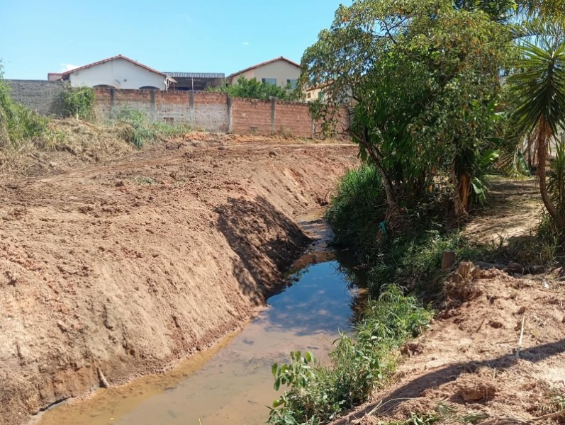 Vista de um canal de água em uma área com vegetação e casas ao fundo, em um dia ensolarado.