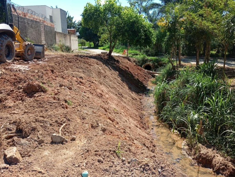 Área de construção com uma máquina escavadeira, margem de um pequeno riacho cercado por vegetação e árvores.