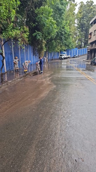 Homens trabalhando na limpeza de uma rua alagada após chuva, com árvores e um muro azul ao fundo.