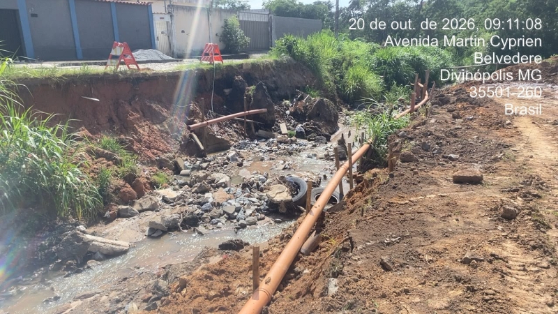 Imagem de uma área em obras na Avenida Martin Cyprien, em Divinópolis, MG, Brasil. Mostra um trecho com erosão, tubos expostos e detritos em um leito de água parada.