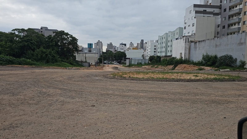 Vista de um terreno vazio com vegetação ao fundo e edifícios na área urbana.