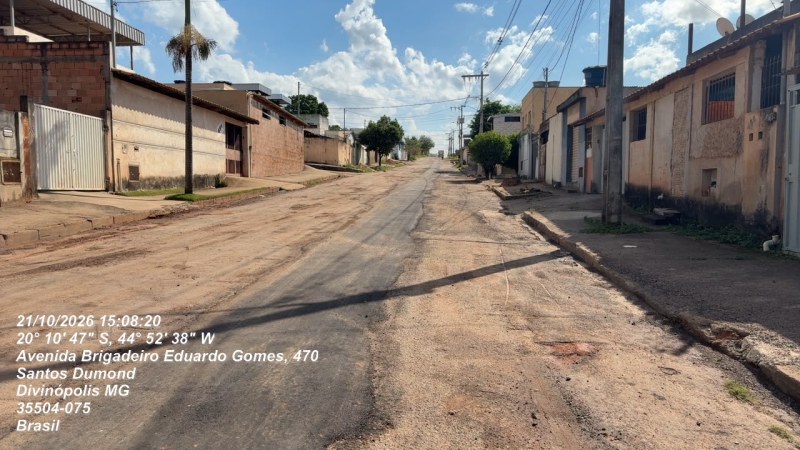 Rua pavimentada com árvore à direita, em um dia ensolarado, com casas ao longo da calçada, localizada em Divinópolis, MG, Brasil.