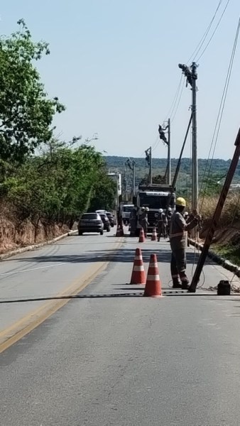 Caminho em uma estrada com cones de sinalização, trabalhadores próximos a um poste e veículos passando.