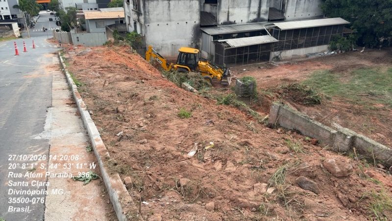 Área em construção com terreno desmatado, onde se vê uma máquina amarela em operação e um espaço vazio atrás de prédios.