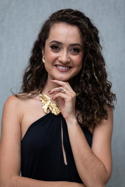 Uma mulher sorridente com cabelo cacheado e adornos de ouro, vestindo um vestido preto, posando com a mão no queixo.