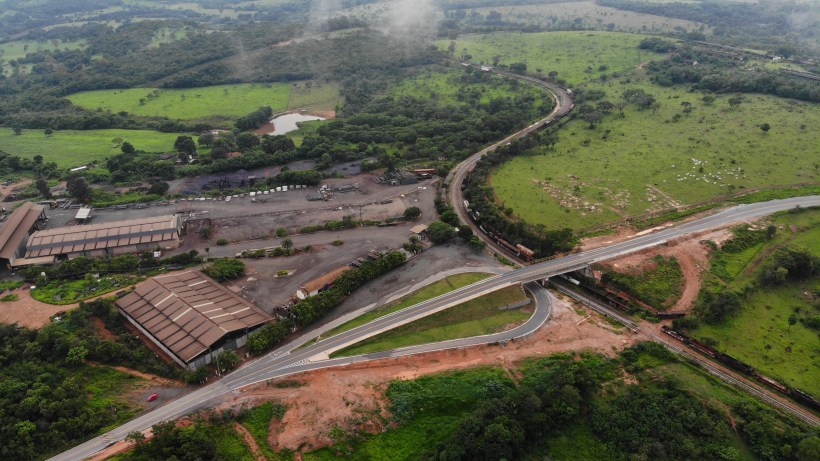 Imagem aérea de uma área rural com estradas sinuosas e um terreno verdejante, incluindo uma área de indústria com edifícios e veículos.