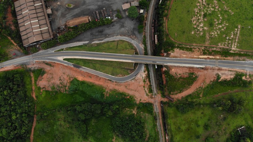 Imagem aérea de uma estrada se curvando sobre uma área rural, com vegetação ao redor e um edifício industrial ao fundo.