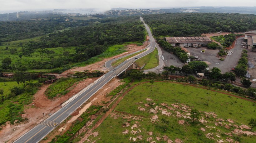 Vista aérea de uma rodovia serpenteando por uma área verde, com terras em construção ao lado e um galpão visível no canto direito.