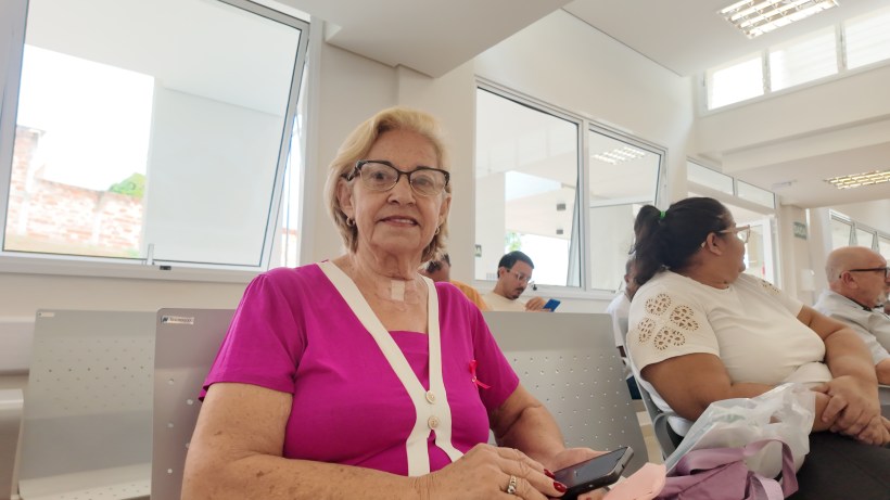 Uma mulher idosa sorrindo em uma sala de espera, vestindo uma blusa rosa e segurando um celular.