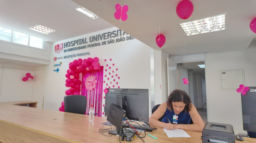 Recepção do Hospital Universitário da Universidade Federal de São João del-Rei, decorada com balões e borboletas rosa. Funcionária trabalhando na mesa.