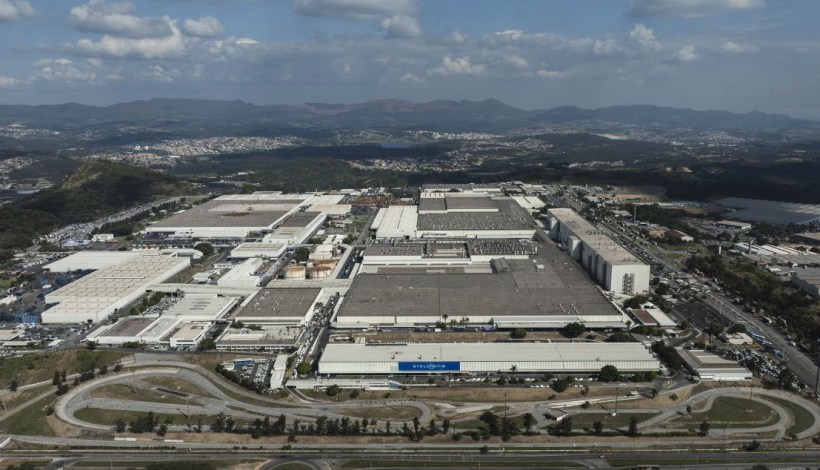 Vista aérea de um complexo industrial, com várias fábricas e armazéns, cercado por montanhas e áreas urbanas ao fundo.