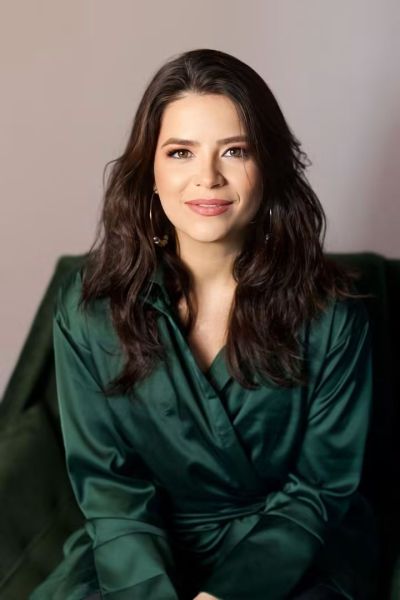 Mulher com cabelo solto e ondulado, usando uma blusa verde, sorrindo em um fundo neutro.
