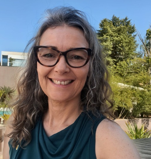Mulher sorridente com cabelo encaracolado e óculos, atrás dela um jardim e um céu azul.